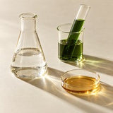 Clear laboratory glassware holding water, a green liquid and a golden liquid in sunlight