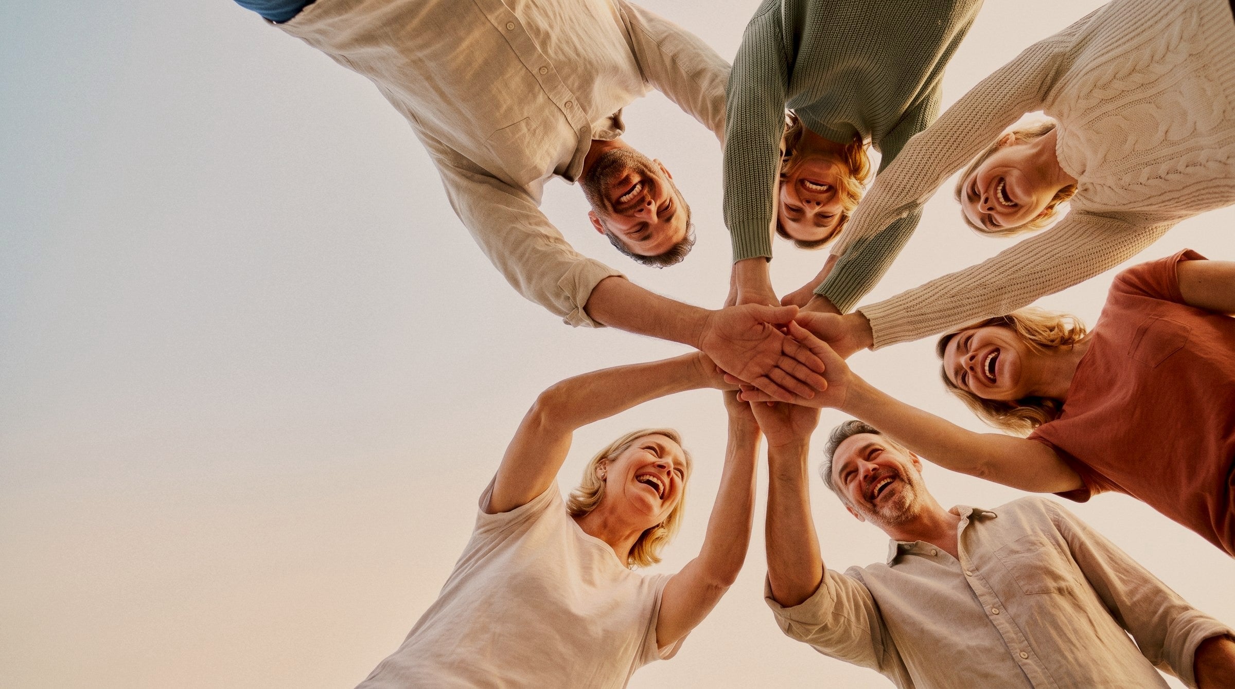 Six friends laughing as they join hands in a circle, seen from below against a pale sky