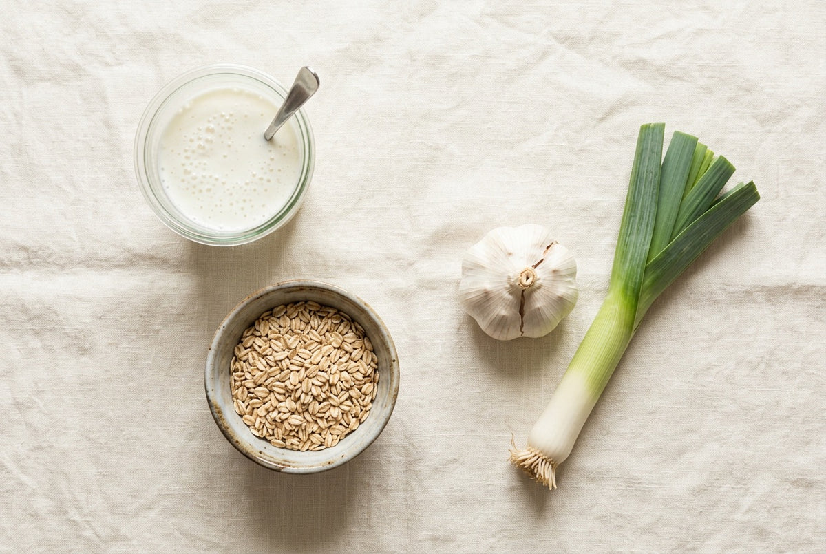 A glass of kefir, a bowl of oats, a garlic bulb and a leek on a cream linen cloth