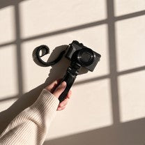 A hand in a cream sleeve holding up a compact camera against a sunlit wall