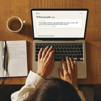 Hands typing on a laptop open at a customer reviews page, beside a notebook and a cup of coffee