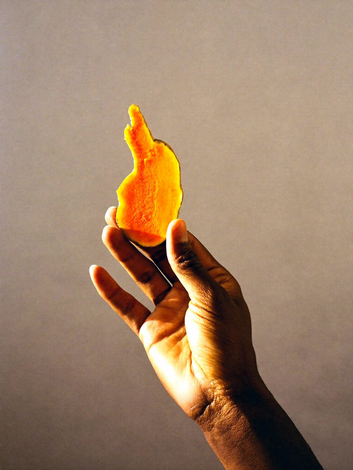 A hand holding up a thin slice of fresh turmeric root against a grey wall