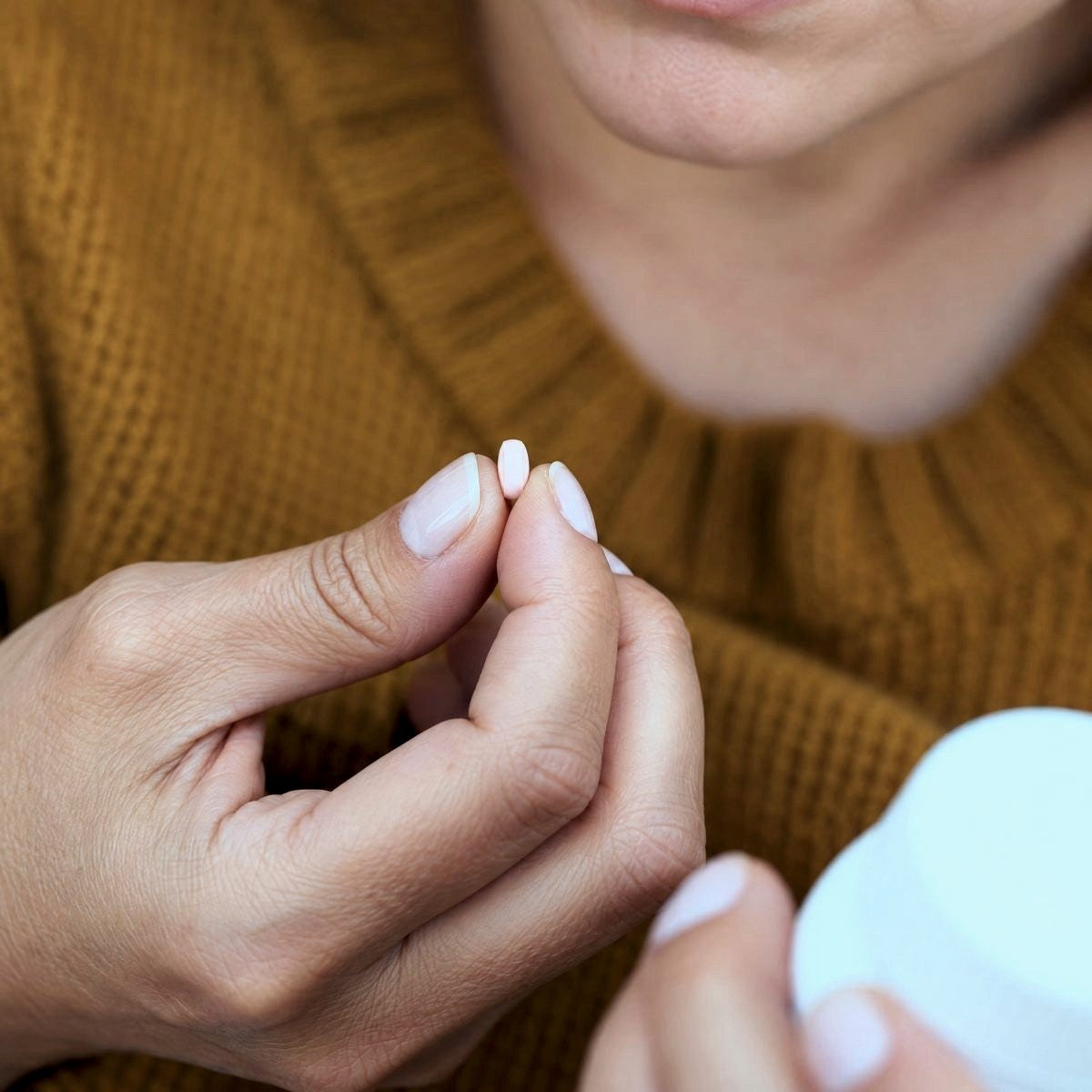 A woman in a mustard jumper holding a small white tablet beside an open pill tub